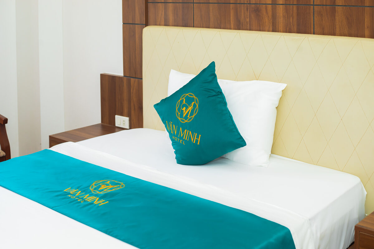 Chi tiết giường với gối và khăn trải màu xanh ngọc thêu logo Văn Minh Hotel màu vàng đồng