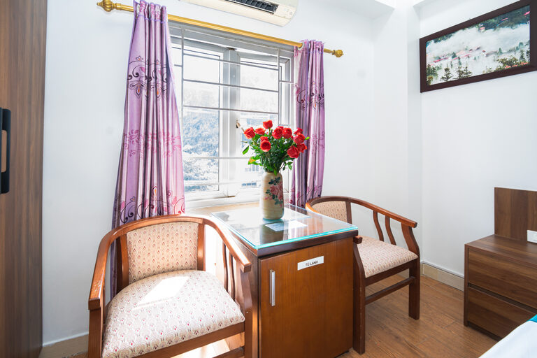 Góc ngồi bên cửa sổ với hai ghế gỗ, bàn kính và bình hoa hồng, nhìn ra đồi núi Bảo Hà
