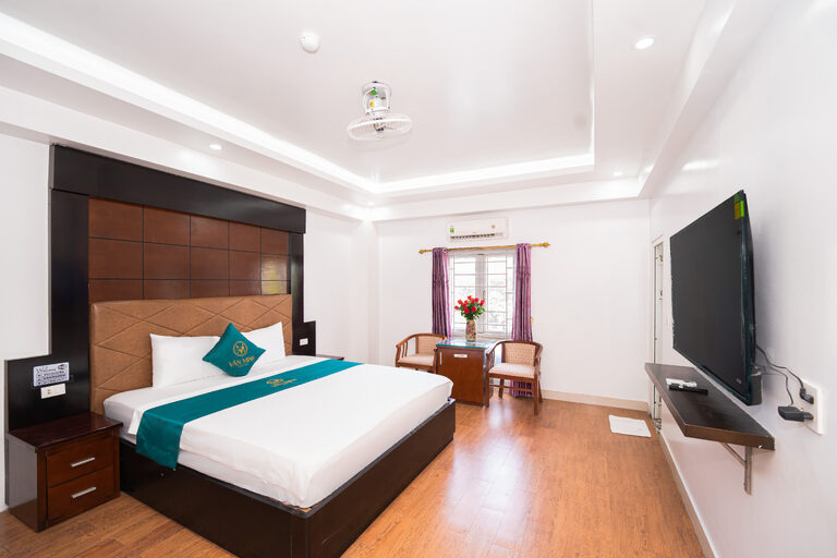 Phòng giường đôi với đầu giường gỗ, góc bàn ghế cạnh cửa sổ và điều hòa