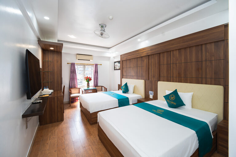 Phòng hai giường đơn với vách đầu giường ốp gỗ, TV màn hình phẳng và góc bàn ghế cạnh cửa sổ
